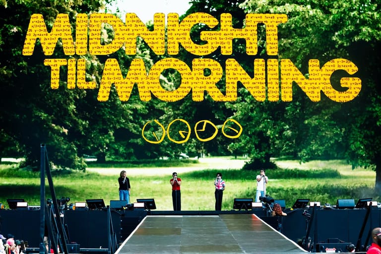 Five band members stand on a stage above a crowd at an outdoor festival venue surrounded by green trees.