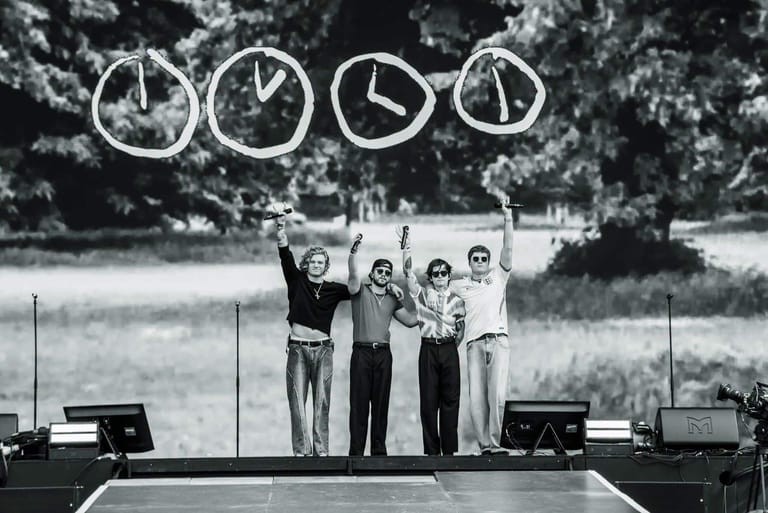 Four performers stand on stage beneath four large suspended clocks in a black and white photograph.