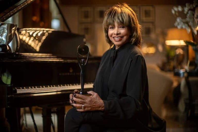 Woman with blonde hair smiling beside a grand piano in a warmly lit studio setting.