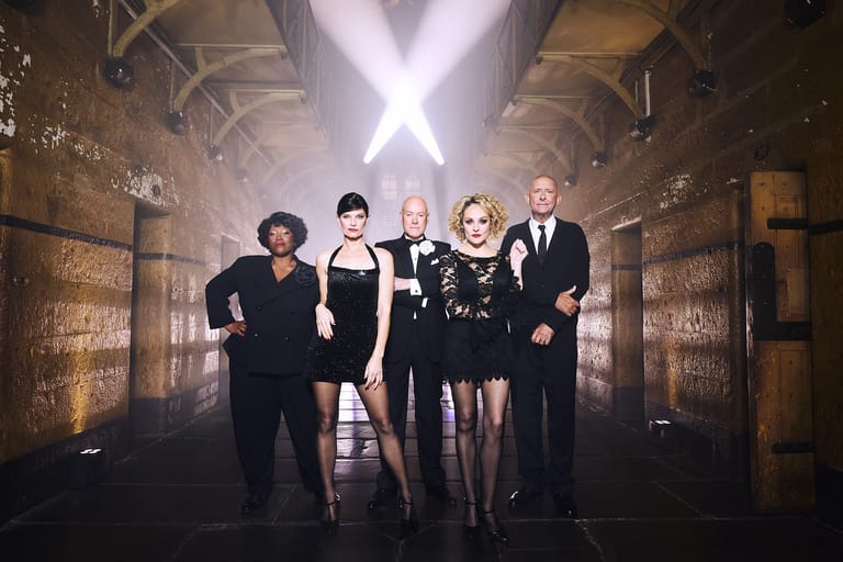 Five performers in black formal attire walk down a theatrical corridor beneath dramatic cross-shaped lighting.