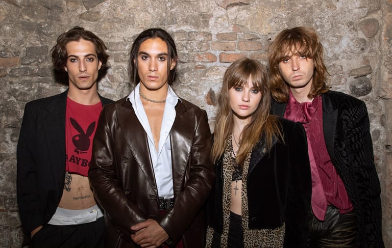 Four members of Maneskin posed against a brick wall in black and burgundy outfits.