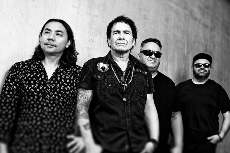 Four band members in black and white, standing against a plain wall with focused expressions.