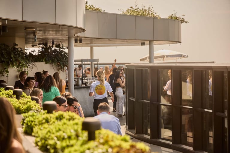 Patrons queue inside a modern café or restaurant with potted plants and warm lighting.