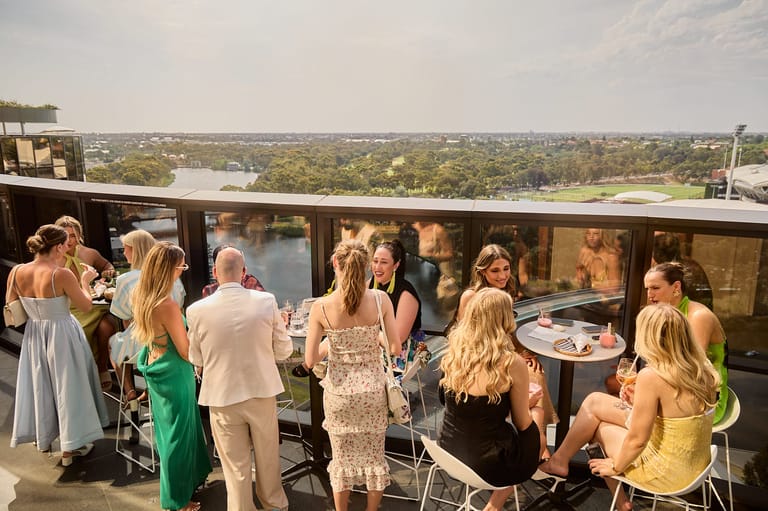 Guests mingle on a high-rise rooftop terrace overlooking Adelaide's parklands and river during an event.