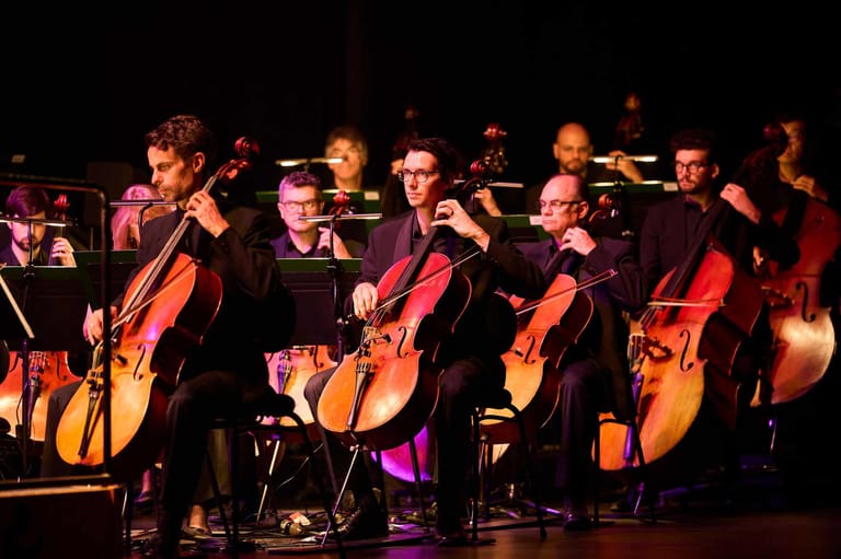 Cellists perform together on stage under warm spotlight lighting during an orchestral concert.