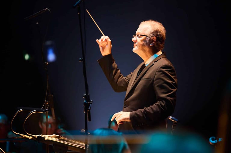 Conductor in brown jacket directing orchestra with baton under blue stage lighting.