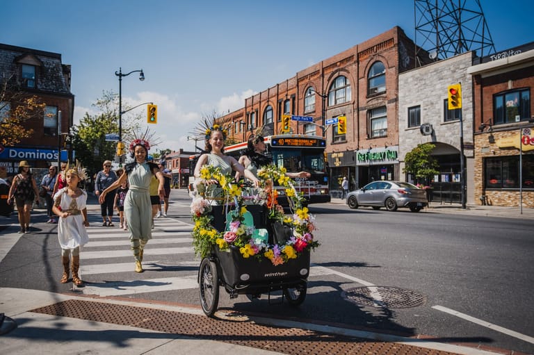 Flower-laden cart crosses pedestrian crossing on sunny street lined with heritage brick buildings.