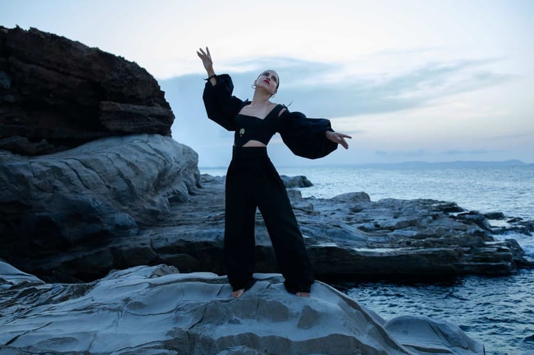 Person in black outfit striking a dramatic pose on rocky coastal outcrop at dusk.