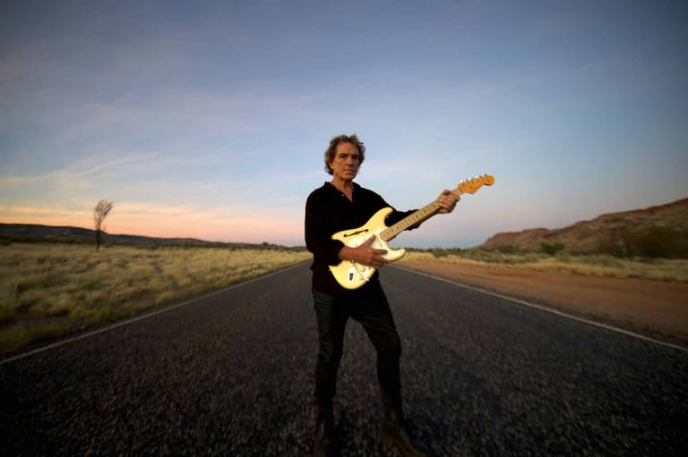 Guitarist in black shirt playing electric guitar on an empty desert highway at sunset.