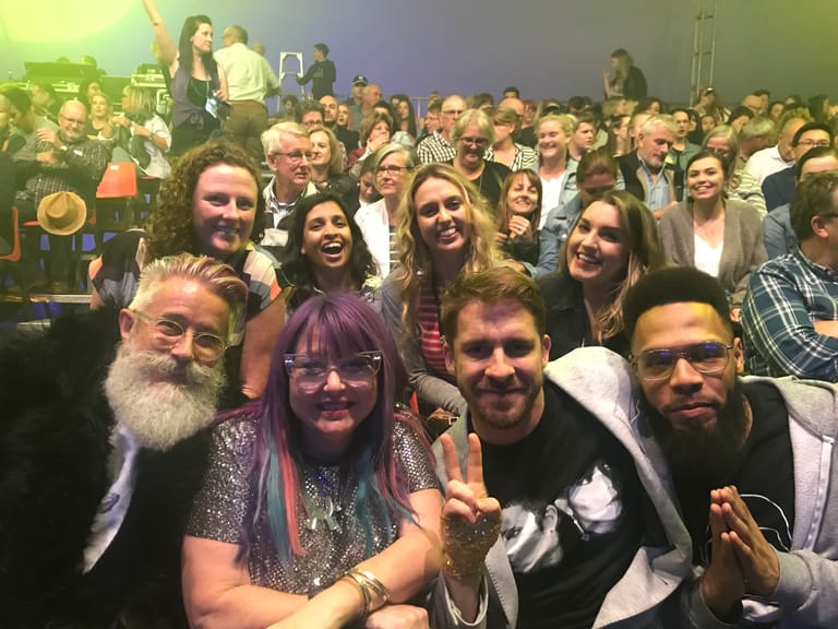 Diverse crowd of people smiling at camera during a lively indoor event or gathering.