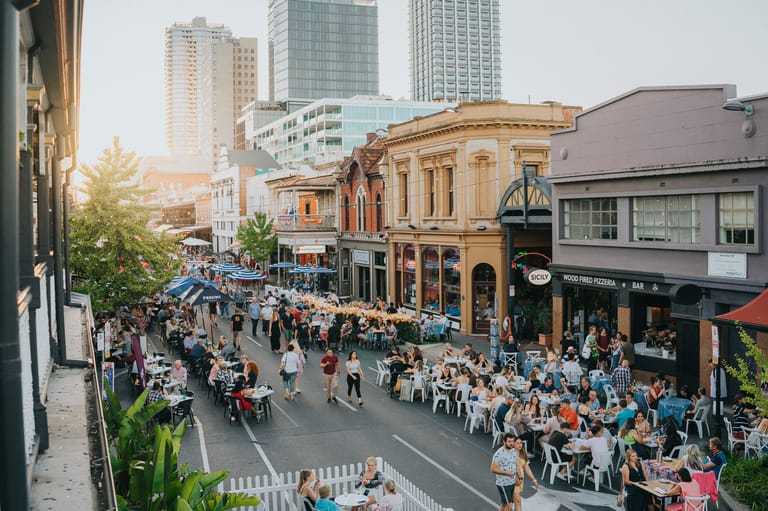 Crowds of people fill a city street beside a grand heritage building with modern high-rises in the background.