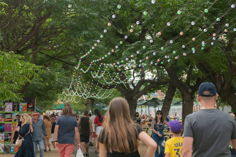 Outdoor festival crowd beneath trees strung with white decorative lights.