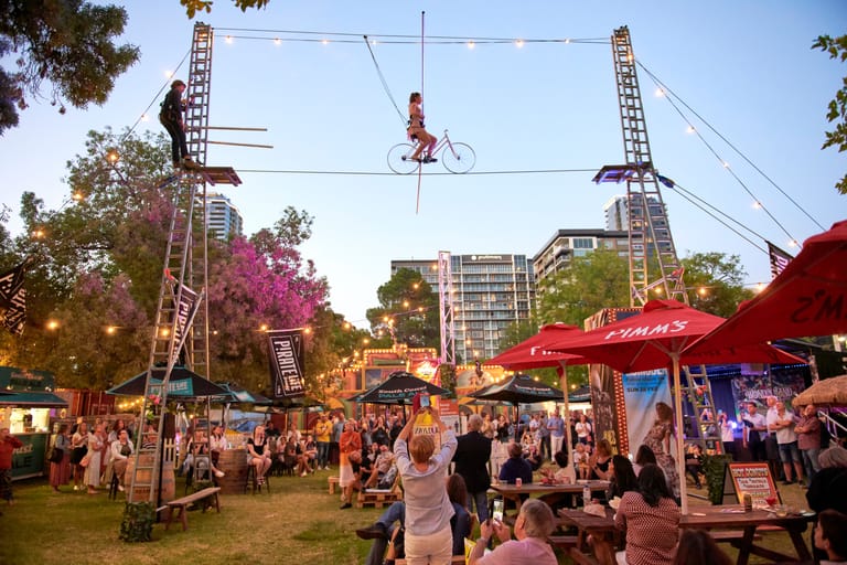 Acrobat suspended on trapeze above outdoor festival crowd beneath tall rigging towers and red umbrellas.