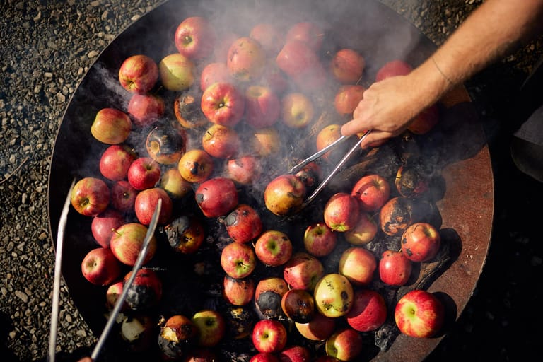 Hand grilling red and yellow apples on a large outdoor flatplate with smoke rising.