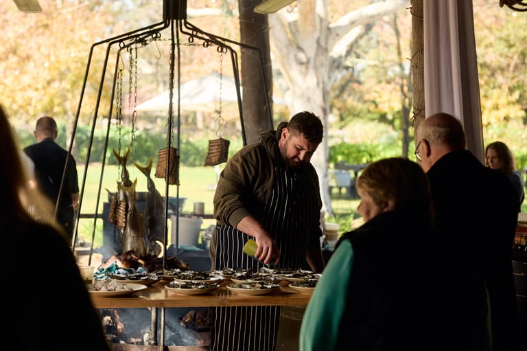 Chef preparing food at a counter while visitors watch through large windows with garden views behind.