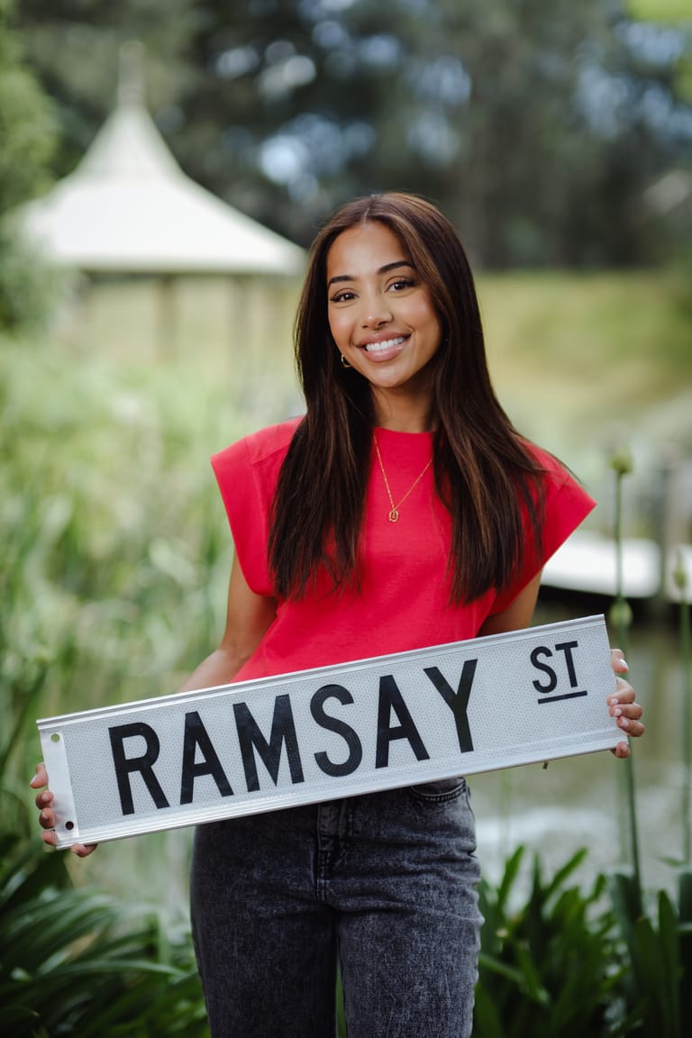 Woman in red shirt holding Ramsay Street sign outdoors with garden backdrop.