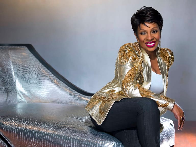 Woman in gold sequined jacket and black trousers posed on silver textured bench against neutral background.