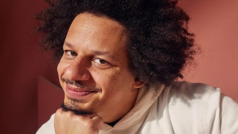 Man with curly hair smiling at the camera against a warm red background.