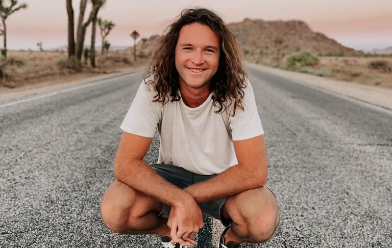 Person with long dark hair crouching on a desert highway, smiling at the camera with mountains visible in the distance.