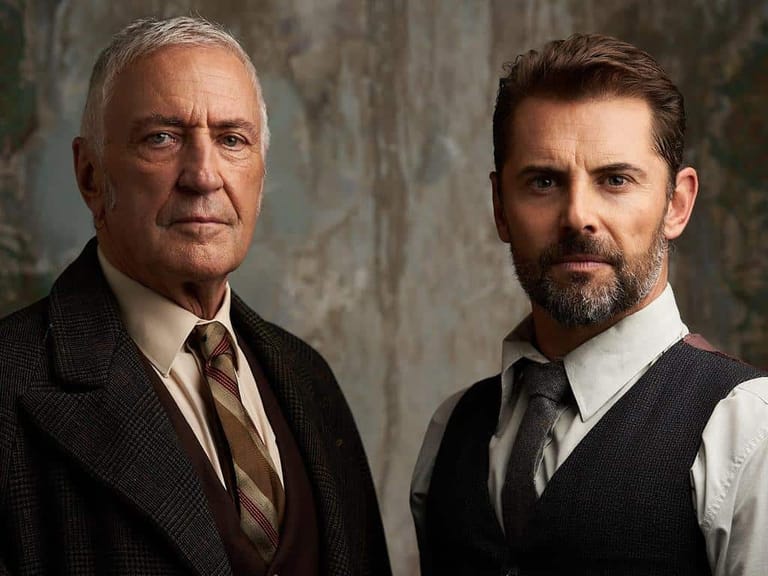 Two men in formal attire stand against a textured grey wall in professional portrait lighting.