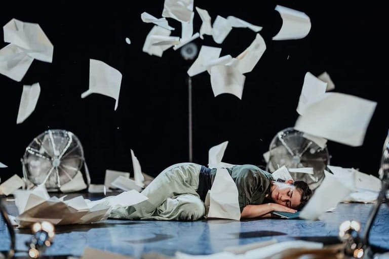 Performer lying on stage surrounded by floating white papers and clear acrylic structures against a dark background.