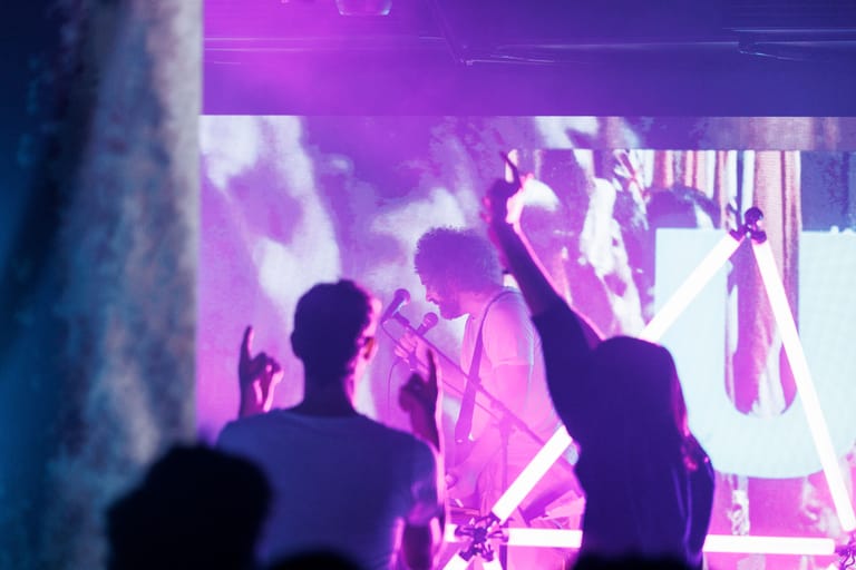 Silhouetted figures dance and raise their arms beneath purple stage lighting at a live performance.