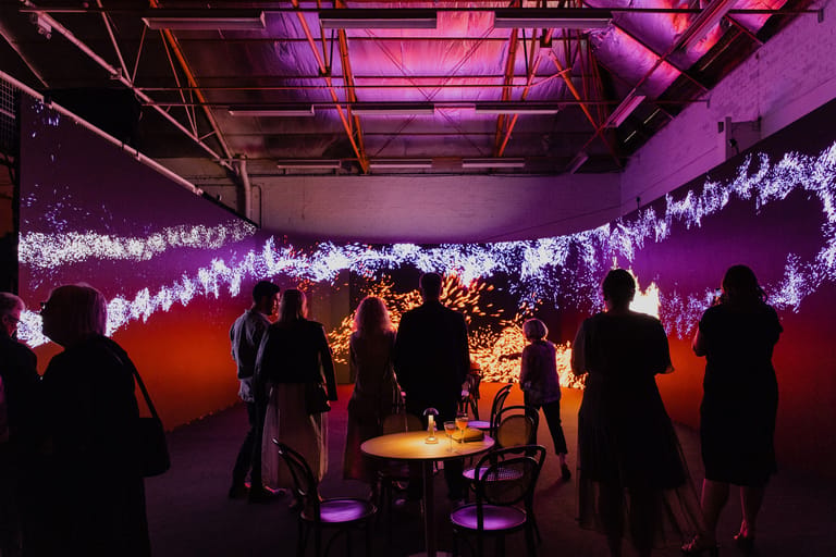 Visitors explore an immersive art installation with glowing purple light trails and warm amber lighting in an industrial gallery space.