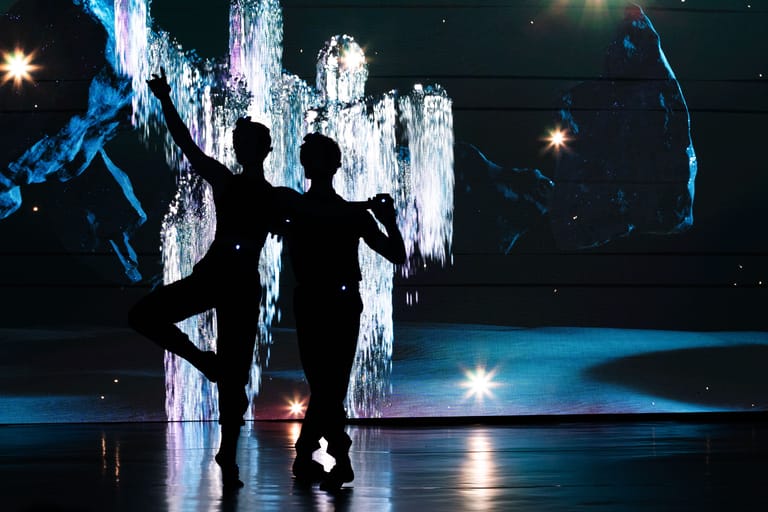 Silhouetted dancer poses against illuminated vertical light installation on dark stage with blue atmospheric lighting.