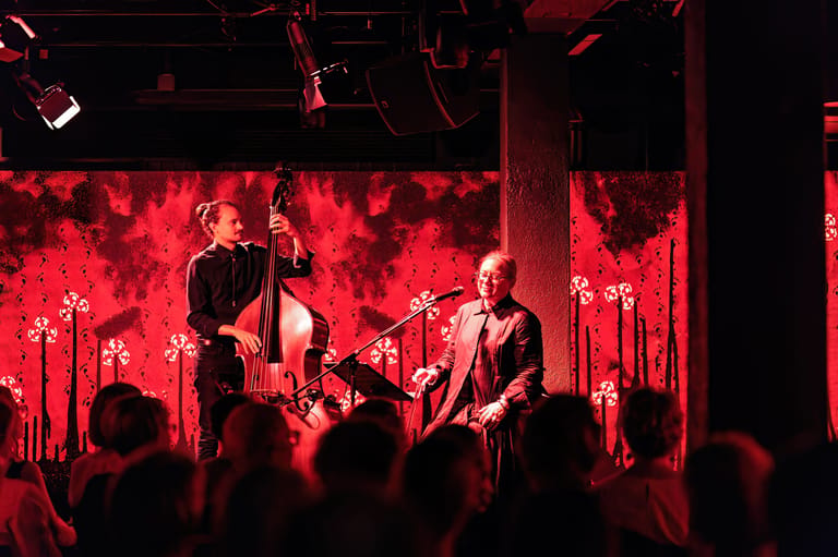 Musicians perform on stage bathed in red light with audience silhouettes in foreground.