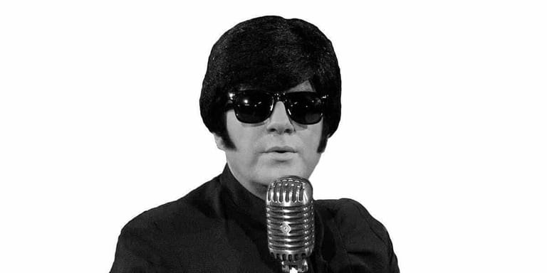 Figure in black sunglasses and dark jacket holding a vintage microphone against white background.