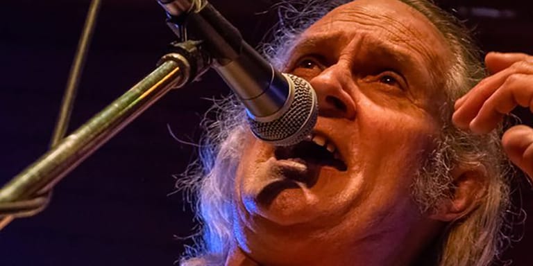 Performer singing intensely at a microphone under blue stage lighting, long grey hair visible.