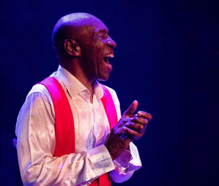 Man in white shirt and red scarf performs on stage under purple spotlights with animated expression.