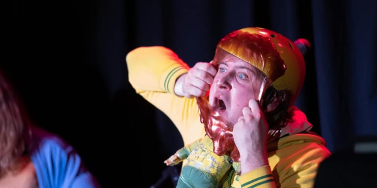 Performer in yellow and red costume singing into microphone under stage lighting.