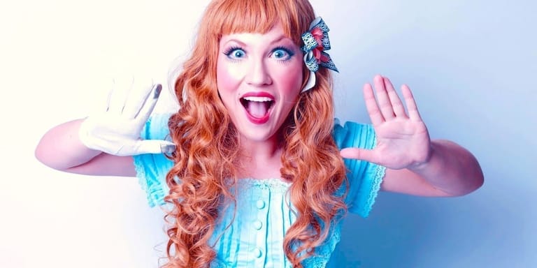 Person with long coral-pink wavy hair and bright makeup wearing turquoise top, gesturing expressively with hands raised.