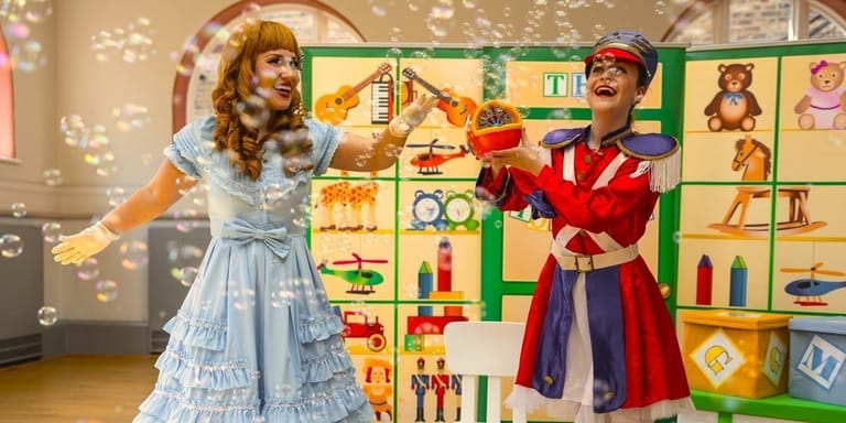Two performers in period costume dance joyfully in a colourful children's theatre set with toy blocks and props.