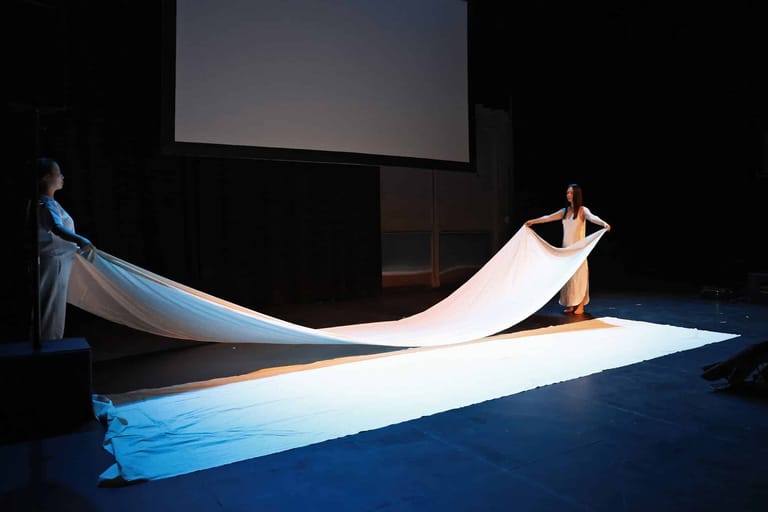 Two performers manipulate flowing blue and white fabric across a darkened stage lit by spotlights.