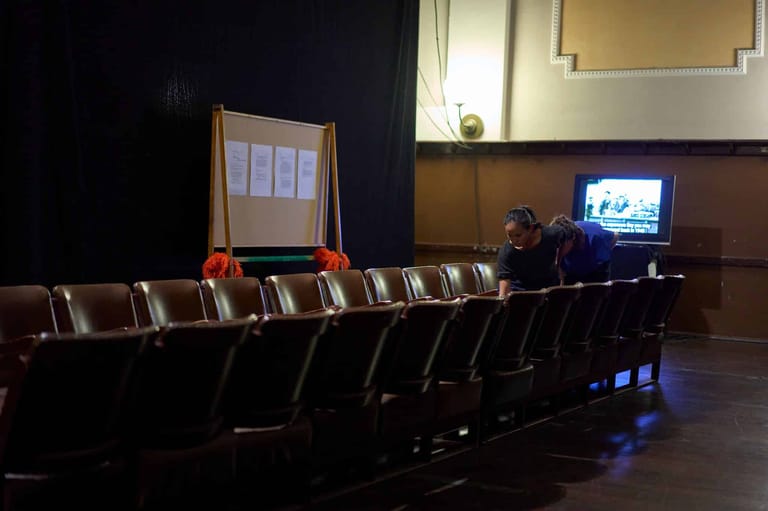 Rows of empty theatre seats face a screen displaying video content in a darkened auditorium.
