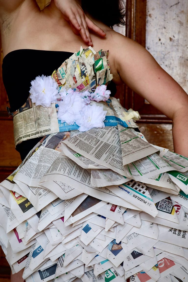 Person holding a large sculptural garment made from layered newspaper and colourful fabric scraps.
