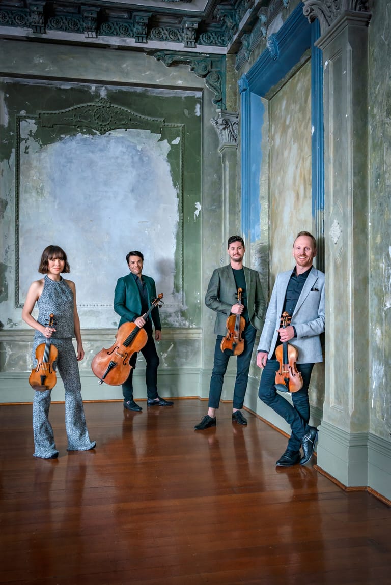 Four string musicians holding violins and cellos pose in a heritage room with blue and green ornate doorways.