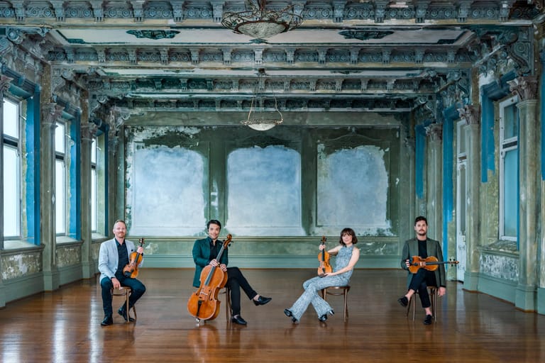Four musicians seated with string instruments in an ornate turquoise hall with high ceilings and large windows.