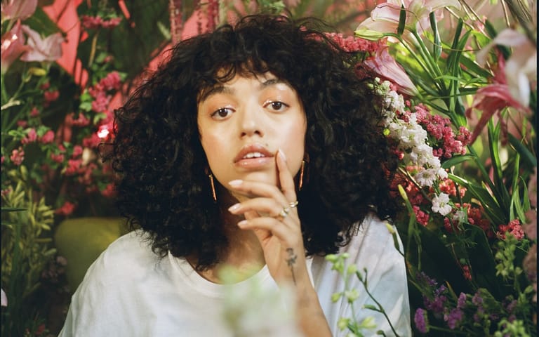 Person with black curly hair and bangs surrounded by pink and white flowers, hand raised thoughtfully to chin.