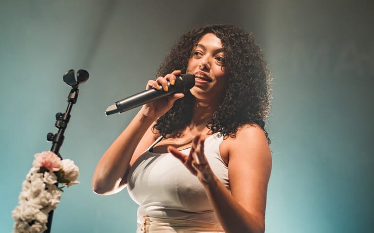 Performer singing into microphone against blue-lit stage backdrop with microphone stand visible.