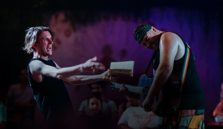 Two performers engage in an intense physical exchange on stage, bathed in purple stage lighting.