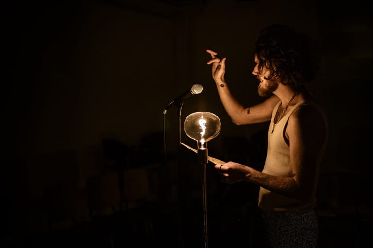 Bearded performer singing at a microphone beneath warm stage lighting against a dark background.