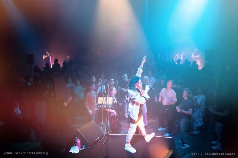 Musician performing on stage in white outfit under blue and red spotlights with crowd gathered around.