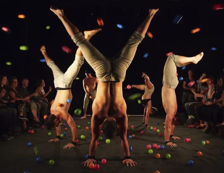 Three acrobats perform handstands surrounded by coloured balls on a darkened stage with audience members seated nearby.