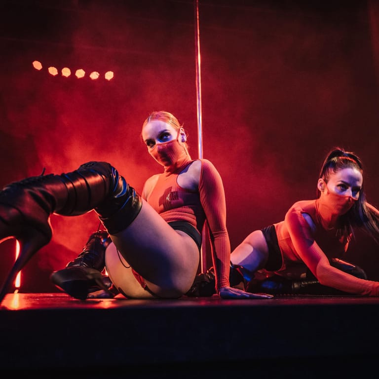 Three performers in athletic wear pose against a pole under dramatic red stage lighting and atmospheric haze.