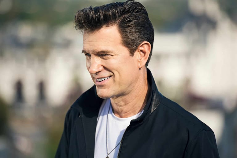Man in black jacket and white shirt smiling outdoors in daylight.