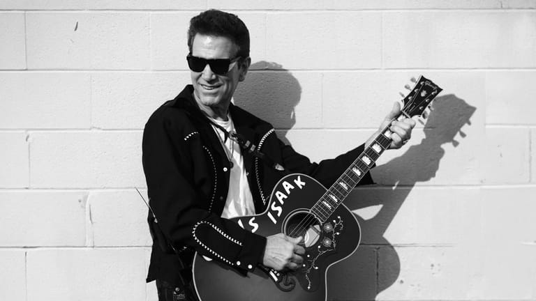 Musician in black leather jacket and sunglasses holding an electric guitar against a white brick wall.
