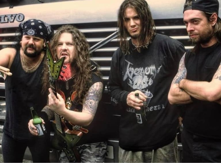 Four musicians in black clothing and long hair stand together in front of a tour bus.